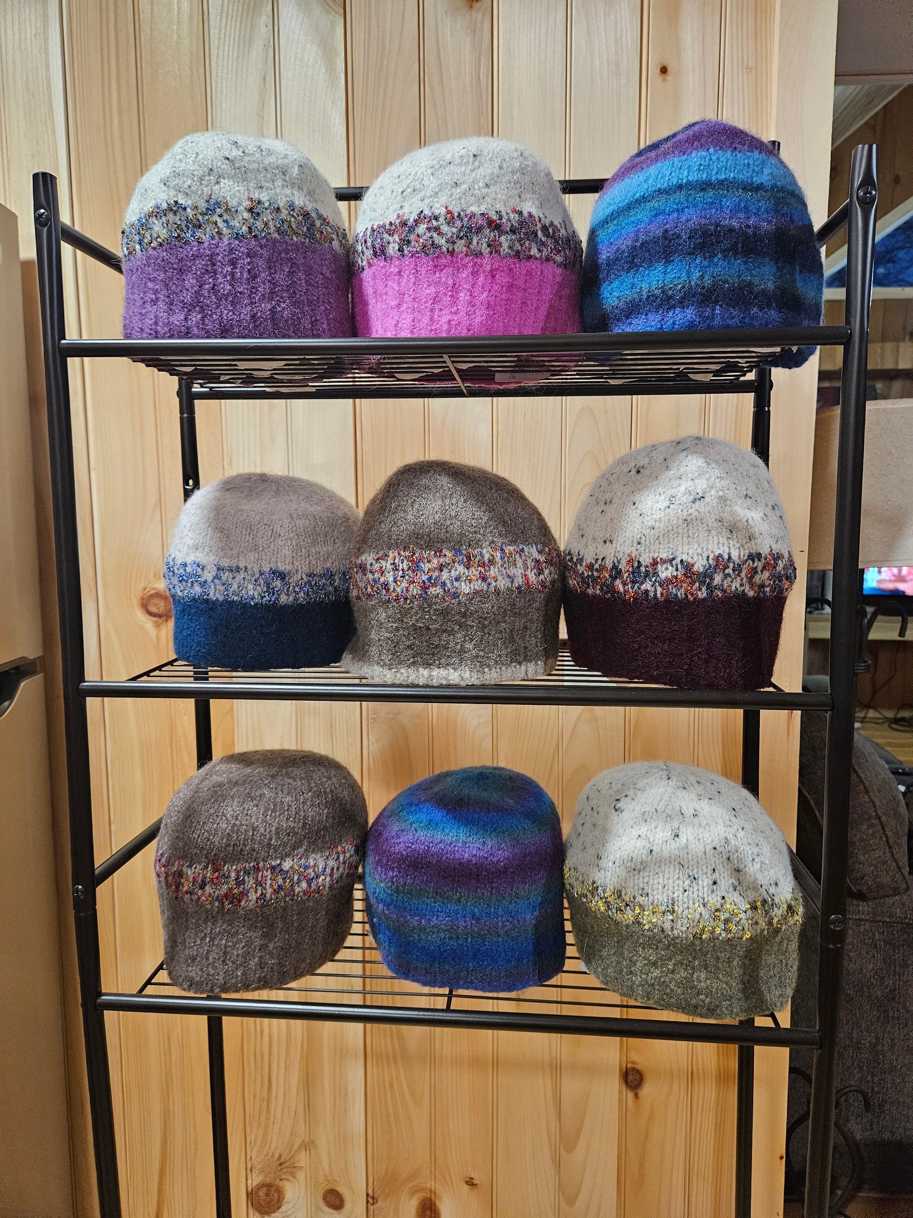 A black wire rack displaying nine hand-felted hats in purple, pink, blue, gray, and burgundy.