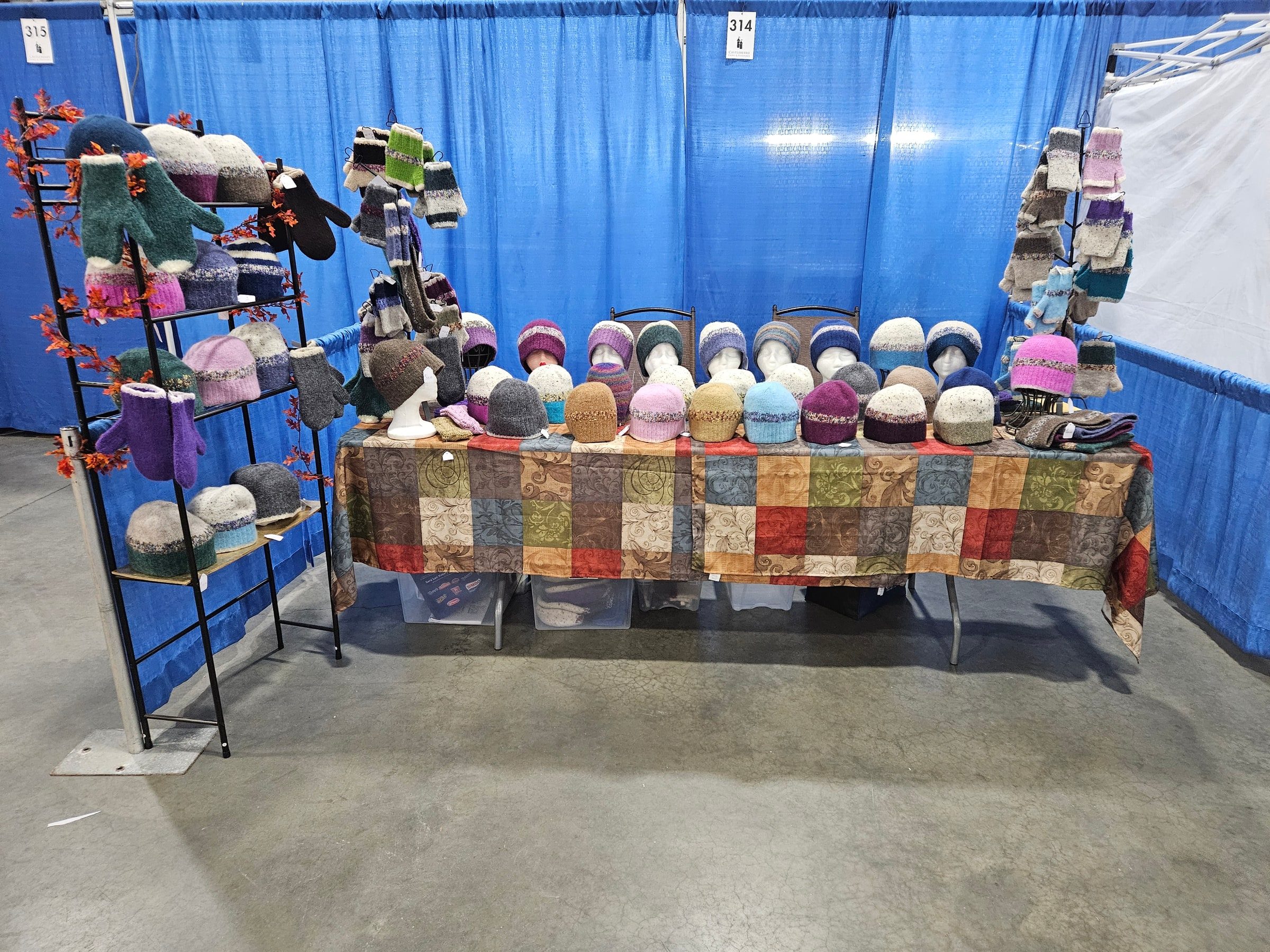 Judy Hella's craft-show booth with hats displayed on racks and a long table under blue curtains.